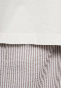 Un tessuto di cotone bianco è stratificato su un tessuto a righe beige, mettendo in mostra una texture liscia e motivi a contrasto. Orlo e cuciture pulite sono visibili.