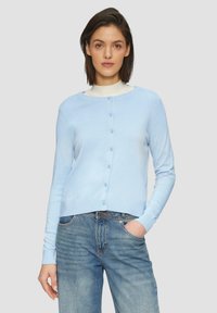 Lichtblauwe cardigan met knoopsluiting, ronde halslijn en lange mouwen. Gemaakt van zacht materiaal, gedragen over een witte top en denim jeans.