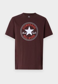 T-shirt en coton marron avec un motif circulaire comportant une étoile blanche, un texte rouge indiquant "CONVERSE ALL STAR" et "Chuck Taylor" dans un cercle intérieur.