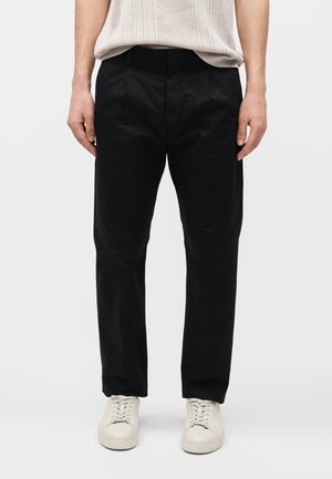 Άνδρας που φοράει μαύρο παντελόνι ραμμένο στα μέτρα και εκρού αθλητικά παπούτσια, στέκεται μπροστά σε ένα απλό φωτεινό φόντο, από τη μέση έως τα πόδια ορατά.