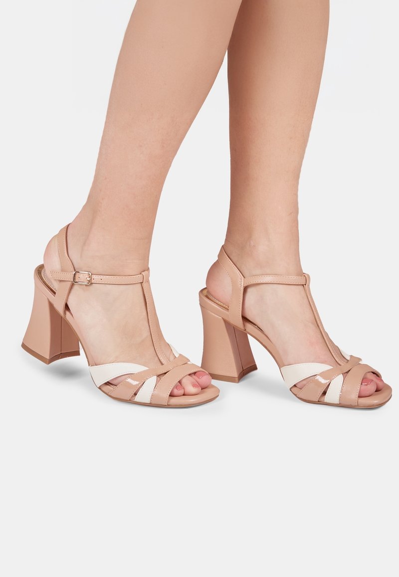 Sandales à talons blocs beiges avec brides en T, présentant une sangle blanche accentuée et une boucle réglable à la cheville.