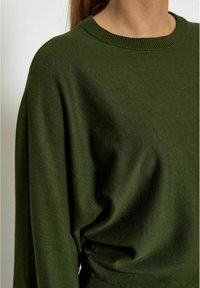 Pull en maille vert olive avec col rond et coupe ample, doté de poignets et d'un ourlet côtelés. Texture douce et légère élasticité visible.