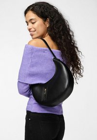 Bolso de cuero negro con forma de media luna, que cuenta con un bolsillo con cremallera y una textura suave, llevado al hombro con un top de punto púrpura.