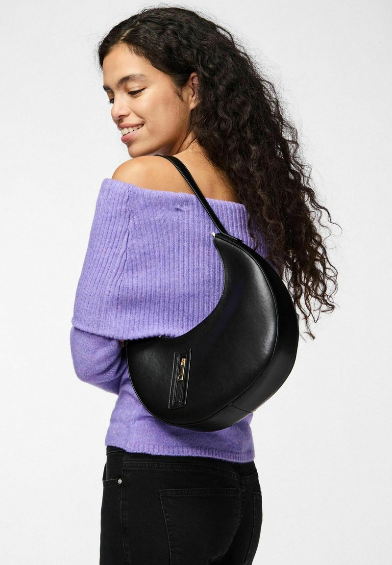 Bolso de cuero negro con forma de media luna, que cuenta con un bolsillo con cremallera y una textura suave, llevado al hombro con un top de punto púrpura.