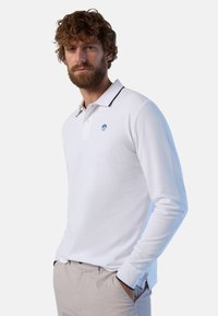 Polo bianco a maniche lunghe con colletto blu navy, caratterizzato da un logo blu sul petto. Il tessuto ha una texture liscia. Indossato con pantaloni beige.