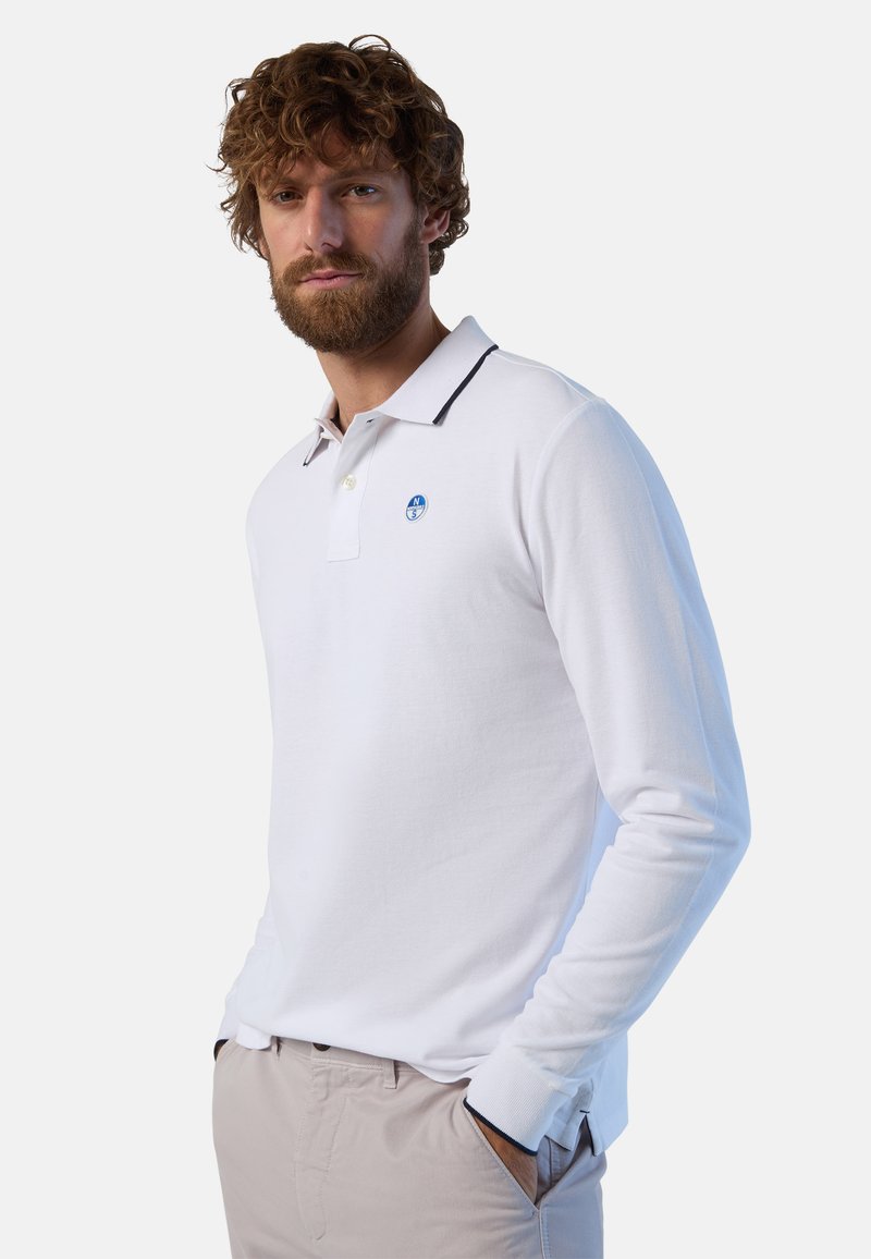 Camisa polo blanca de manga larga con cuello azul marino, que presenta un logo azul en el pecho. La tela tiene una textura suave. Se lleva con pantalones beige.