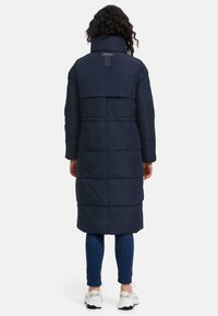 Marinblå lång pufferjacka med hög krage, med en quiltad design och en märkesetikett på baksidan, bärs över blå jeans och sneakers.