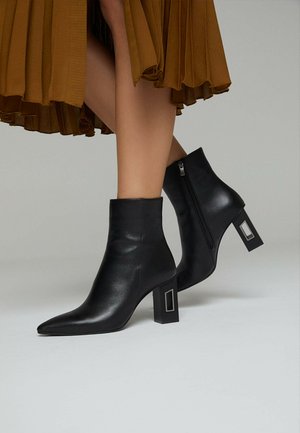 Botas de cuero negro hasta el tobillo con puntas afiladas y tacones cuadrados que presentan acentos metálicos, además de una cremallera lateral para facilitar su uso. Falda de tela texturizada visible.