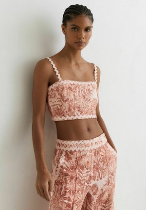 Femeie purtând un top crop fără mâneci și pantaloni cu talie înaltă asortați, cu imprimeu botanic roz și rugină și margine ondulată, stând în picioare lângă un perete alb.