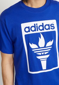 Blue cotton t-shirt featuring a large white Adidas logo and torch graphic on the front. Crew neck with short sleeves.