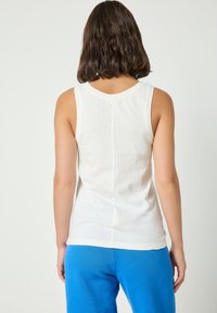 Frau mit braunen schulterlangen Haaren, die ein ärmelloses weißes Oberteil und eine blaue Hose trägt, steht mit dem Rücken zur Kamera.