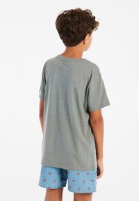 Camiseta de algodón gris de manga corta, vista desde la parte de atrás. Combinada con pantalones cortos azules con pequeños patrones de palmeras rojas.