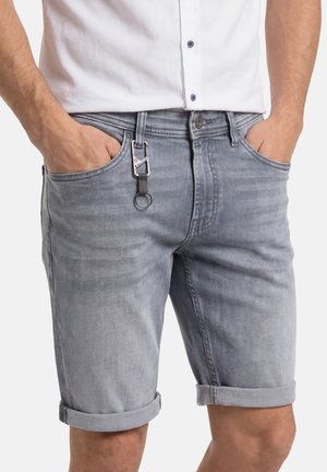 Mann trägt hellgraue Jeansshorts mit umgeschlagenen Säumen, ein weißes Hemd, Hände in den Taschen und einen Schlüsselanhänger an der vorderen Gürtelschlaufe.