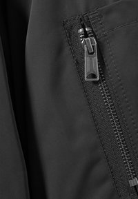 Κοντινή λήψη φερμουάρ μαύρου υφασμάτινου σακακιού με μεταλλικό έλκτη, μερικώς κλεισμένο, που δείχνει λεπτομερείς ραφές και υφή.