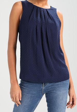 Granatowa teksturowana bluzka bez rękawów z plisami z przodu, noszona z niebieskimi dżinsami na osobie.