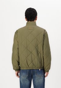 Quiltet olivengrøn jakke med høj krave, elastiske manchetter og kant. Har et lille rødt logo på ryggen. Letvægtsstof.