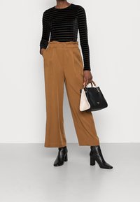 Schwarz samtene Langarmbluse mit vertikalen Streifen, sandfarbene weite Hosen mit Gürtel und eine zweifarbige Handtasche in Schwarz und Creme. Schwarze Absatzstiefeletten.