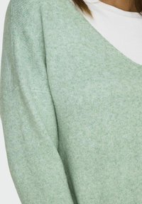 Svetlo zelena pletena pulover s V-izrezom. Mehek material, dolgi rokavi in nekoliko ohlapen kroj. Prevladuje čez belo majico.