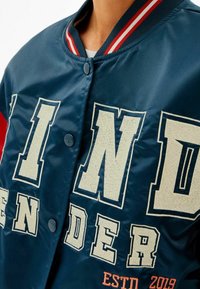 Marinblå bomberjacka med rödvit randig krage. Har stora off-white broderade bokstäver och knappstängning. Mjukt, glansigt tyg.