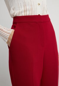 Rote maßgeschneiderte Hosen mit glatter Textur, Seitentaschen und einem strukturierten Bund, kombiniert mit einem weißen, gefalteten Langarmhemd.