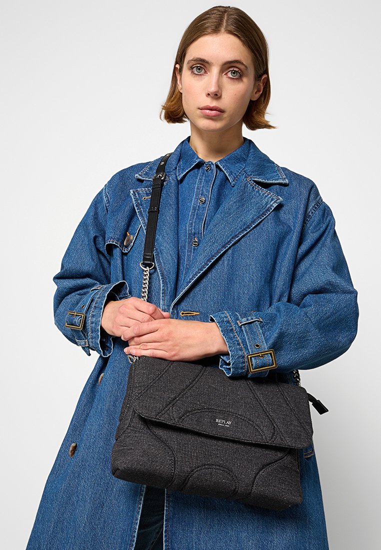 Borsa in tessuto nero con texture, dotata di una patta pieghevole, caratterizzata da un motivo sottile e una tracolla a catena, tenuta da una persona con una giacca di denim.