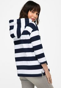 Haut à capuche rayé bleu marine et blanc avec une poche frontale, des longues manches et des fentes sur les côtés. Le tissu semble doux et légèrement texturé.