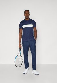 Maglietta atletica blu con striscia orizzontale bianca, abbinata a pantaloni da jogging blu. Scarpe da ginnastica bianche. In mano tiene una racchetta da tennis.