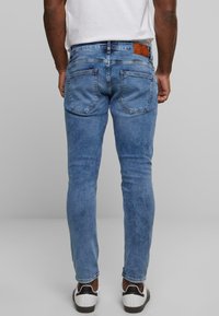 Világoskék farmernadrág, slim fit szabással, két hátsó zsebbel és barna bőr címkével a derekán, fehér sportcipővel viselve.