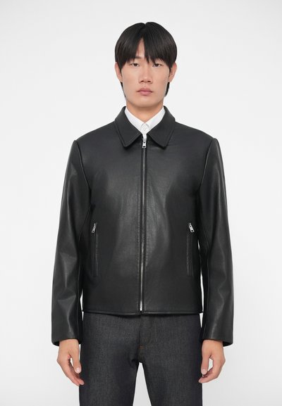 Giacca in pelle nera con colletto piccolo, chiusura frontale con zip e due tasche laterali con zip. Il modello la indossa sopra una camicia bianca e jeans scuri.