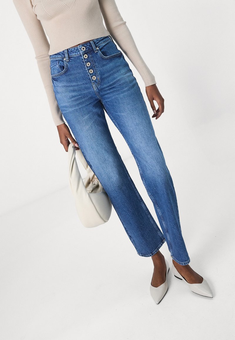 Hochgeschnittene blaue Jeans mit geradem Bein, Fünf-Knopf-Verschluss und vorderen Taschen. Kombiniert mit einem hellbeigen Oberteil und einer weißen Tasche.