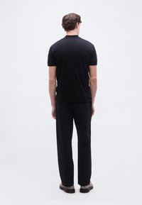 Άνδρας που στέκεται με την πλάτη προς τα εμπρός, φορώντας μαύρο μπλουζάκι με κοντά μανίκια, μαύρο παντελόνι και ανοιχτόχρωμα παπούτσια σε λευκό, απλό φόντο.