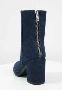 Bottine en denim avec un talon bloc haut, dotée d'une fermeture éclair sur le côté et d'une texture lisse. Couleur bleu foncé avec un détail de couture.
