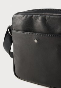 Μαύρη δερμάτινη τσάντα ώμου με λεία υφή, που διαθέτει κλείσιμο με φερμουάρ, μπροστινή τσέπη και ασημί μεταλλική λεπτομέρεια.