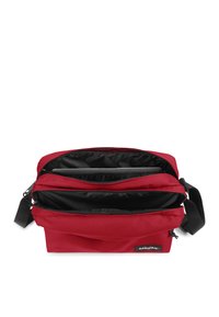 Röd tyg axelväska med flera dragkedjefack, svart foder, justerbar rem och Eastpak-logotyp på sidan.