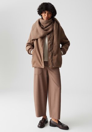 Giubbotto bomber marrone con polsini a coste, indossato sopra un top beige. Pantaloni marroni a gamba larga, abbinati a mocassini in pelle marrone scuro. Sciarpa testurizzata.