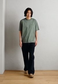 T-shirt vert olive à manches courtes avec une poche poitrine, associé à un pantalon large noir et des chaussures beiges, sur fond neutre.