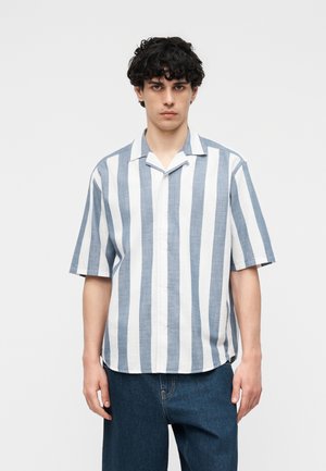 Ung mand med krøllet hår iført en kortærmet, knappet skjorte med brede, lodrette hvide og lyseblå striber og mørke jeans.