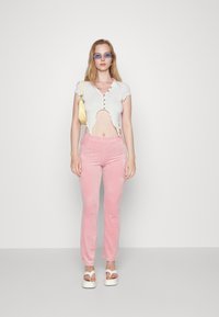 Top branco cropped com bordas rendadas e botões, combinado com calças de veludo rosa claro. Sandálias brancas de plataforma e uma pequena bolsa amarela realçam o visual.