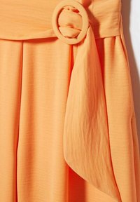 Tissu orange à texture lisse. Comprend une ceinture avec une boucle ronde et une queue drapée, créant un design plat et fluide.