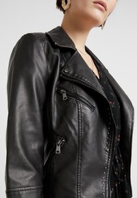 Giacca da motociclista in pelle nera con accessori in metallo argentato, tasche con zip e dettagli borchiati. Texture morbida e vestibilità su misura.