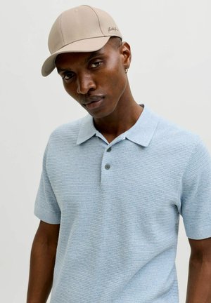 Man draagt een beige pet, kleine oorring en een lichtblauw polohemd met korte mouwen en een gestructureerde stof, tegen een effen achtergrond.