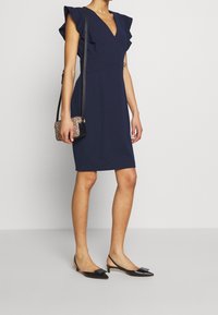 Robe marine ajustée avec des manches volantées et un décolleté en V profond. Associée à des chaussures plates noires et un petit sac à main à motifs.