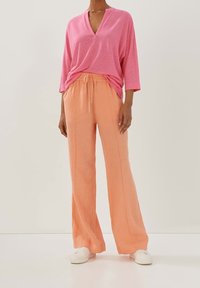 Rosa V-ringad skjorta med avslappnad passform, tillsammans med löst sittande, persikofärgade linbyxor och vita sneakers, mot en neutral bakgrund.