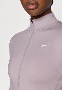 Αθλητι κό τοπ σε ανοιχτό μωβ με ψηλό γιακά και μισό φερμουάρ. Διαθέτει λευκό λογότυπο Nike στο στήθος και λείο, ελαστικό ύφασμα.