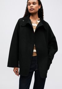 Kvinde iført en sort oversized frakke over en tan- og hvidstribet zip-sweater med mørke jeans, stående mod en lys baggrund.