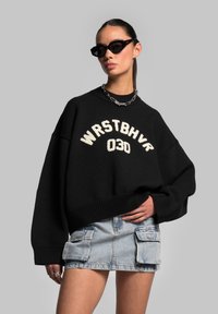 Black oversized knitted sweater with cream embroidery reading "WRSTBVR 030," paired with a light denim cargo mini skirt and sunglasses.