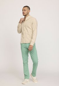 Chemise beige à manches longues en tissu léger, col mandarin et boutons en bois, associée à un pantalon vert menthe et des baskets beige clair.