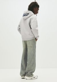 Grijze hoodie met blauw logo op de achterkant, gecombineerd met wijde gestreepte spijkerbroek en witte sneakers.