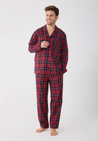 Man smiling, wearing red and navy blue plaid long-sleeve pajama top and matching pants, standing barefoot on white background.