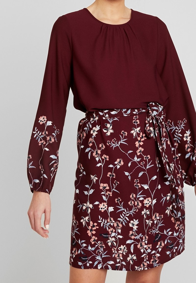 Femme portant une robe longue bordeaux à manches longues avec des motifs floraux sur les manches et la jupe, taille nouée et encolure arrondie.